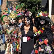 A group in elaborate Dia de los Muertos costumes, face paint, and towering feathered headdresses at the festival