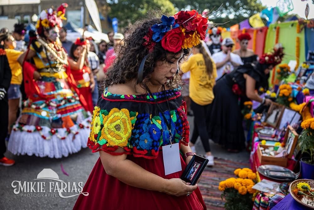 A woman in a traditional embroidered blouse and red skirt with a flower crown, holding a framed photo of a loved one at the community ofrenda